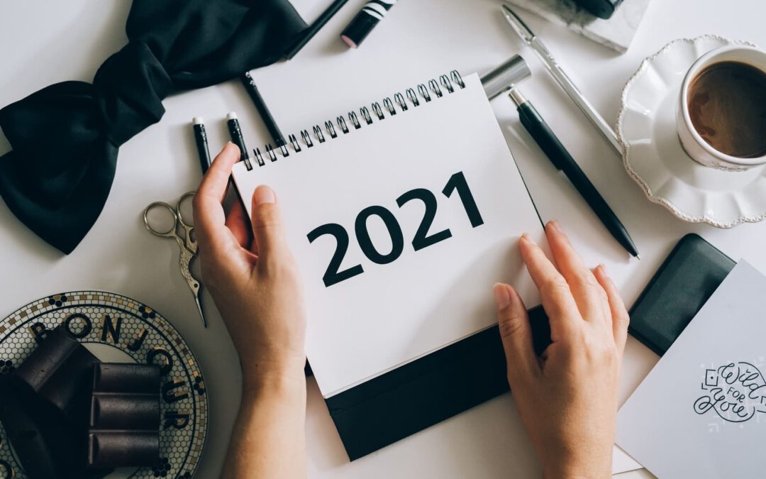 2 Hände halten einen Abreißkalender mit dem Jahr 2021 über einem Tisch, auf dem stilvolle schwarz-weiße Dekoelemente drapiert sind, wie eine Schleife, Kugelschreiber und eine Schere. Auch eine Tasse Kaffe steht rechts oben in der Ecke.