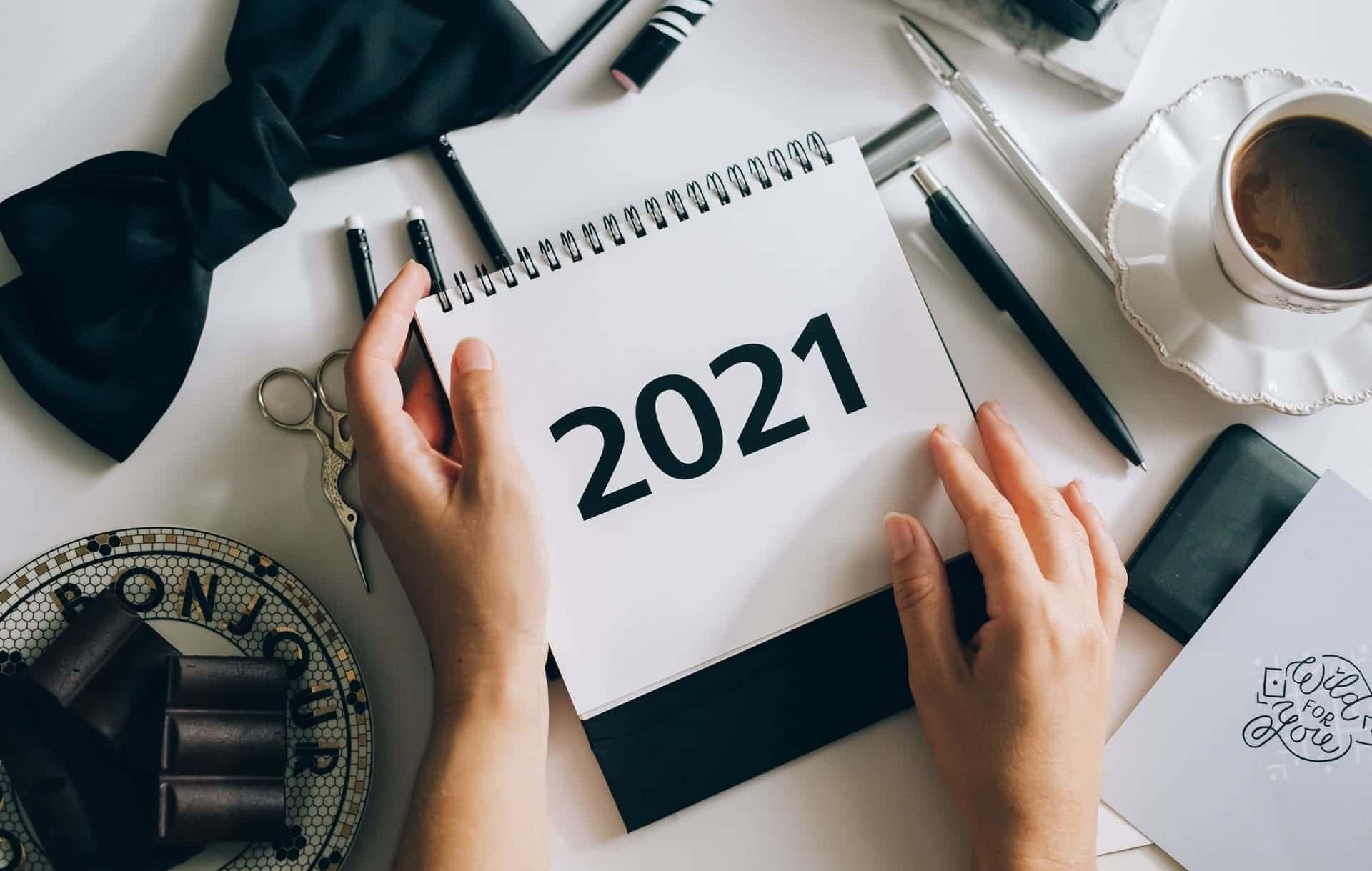 2021 Neues Jahr Vorsätze 2 Hände halten einen Abreißkalender mit dem Jahr 2021 über einem Tisch, auf dem stilvolle schwarz-weiße Dekoelemente drapiert sind, wie eine Schleife, Kugelschreiber und eine Schere. Auch eine Tasse Kaffe steht rechts oben in der Ecke.