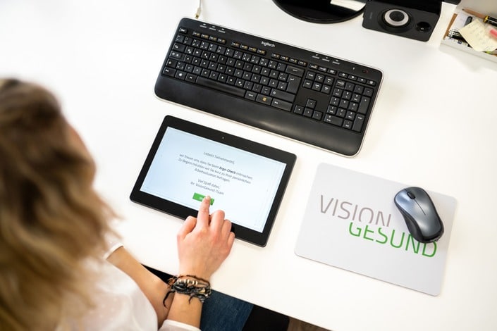 Im Mittelpunkt des Bildes liegt ein schwarzes Tablet, vor einer Computertastatur. Rechts daneben befindet sich eine Maus auf einem VisionGesund-MousePad. Der Kopf einer blonden Frau ist in der linken Bildhälfte zu erkennen: Sie tippt auf dem Tablet vor sich und trägt eine weiße Bluse.