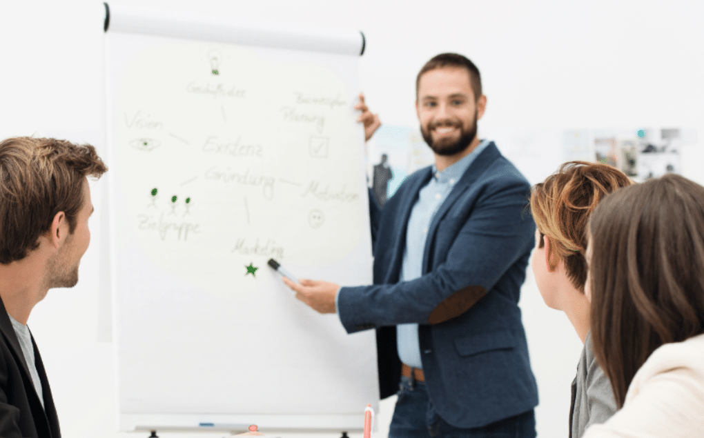 Gesundes Führen Mehrere Personen sitzen in einem Büro-Workshop, eine Person erklärt Inhalte an einem Flipchart. Das Bild zeigt Betriebliche Gesundheitsförderung durch gemeinsames Lernen und Austausch am Arbeitsplatz.