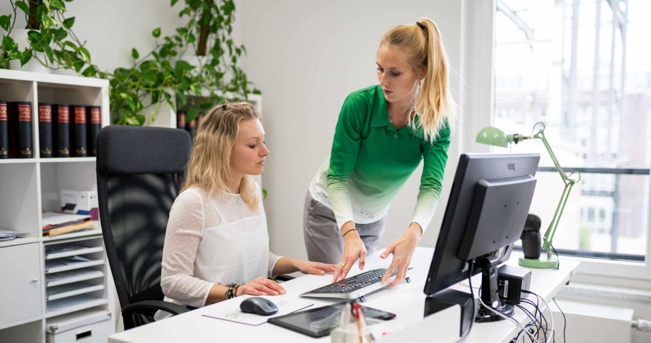 Zwei Personen sitzen an einem modernen Büroarbeitsplatz mit Monitoren und besprechen ergonomische Anpassungen. Der Raum wirkt hell und funktional eingerichtet.
