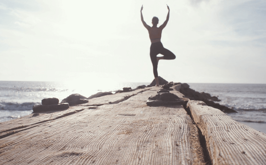 Resilienz Person praktiziert Yoga in der Baumhaltung auf einem hölzernen Steg am Meer. Silhouette vor hellem Himmel, Fokus auf Achtsamkeit, Balance, Stressreduktion und ganzheitliche Gesundheitsförderung in der Natur.