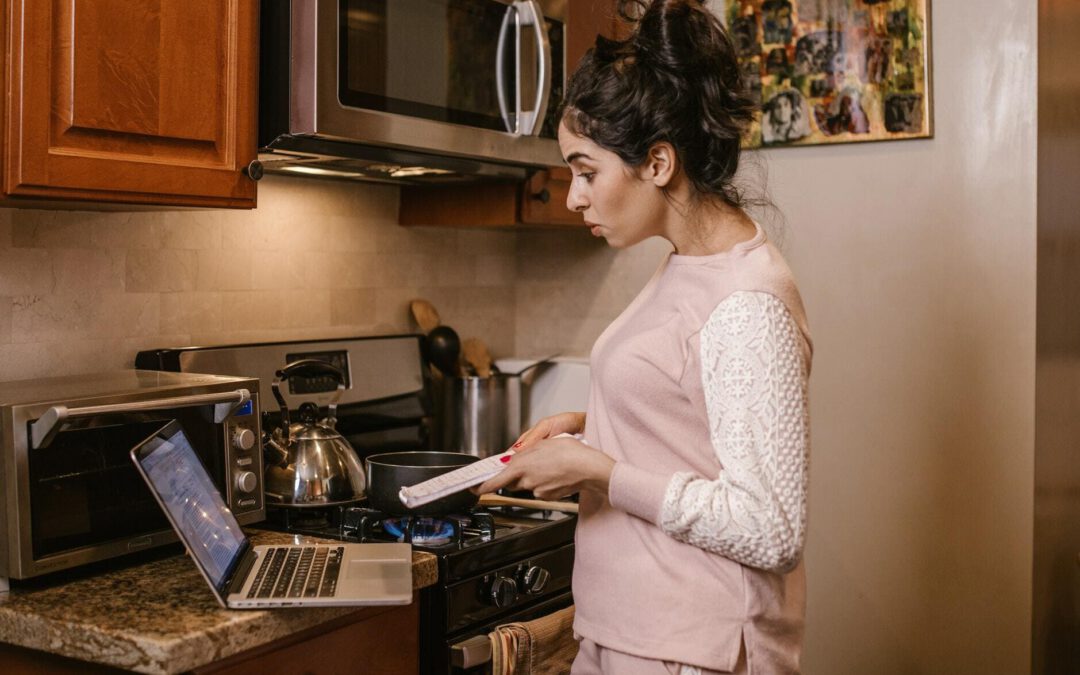 Eine Frau im pastellfarbenen Hausanzug steht vor dem Herd und schaut konzentriert in ihren Laptop auf der Arbeitsplatte. In der Hand hält sie einige Unterlagen.