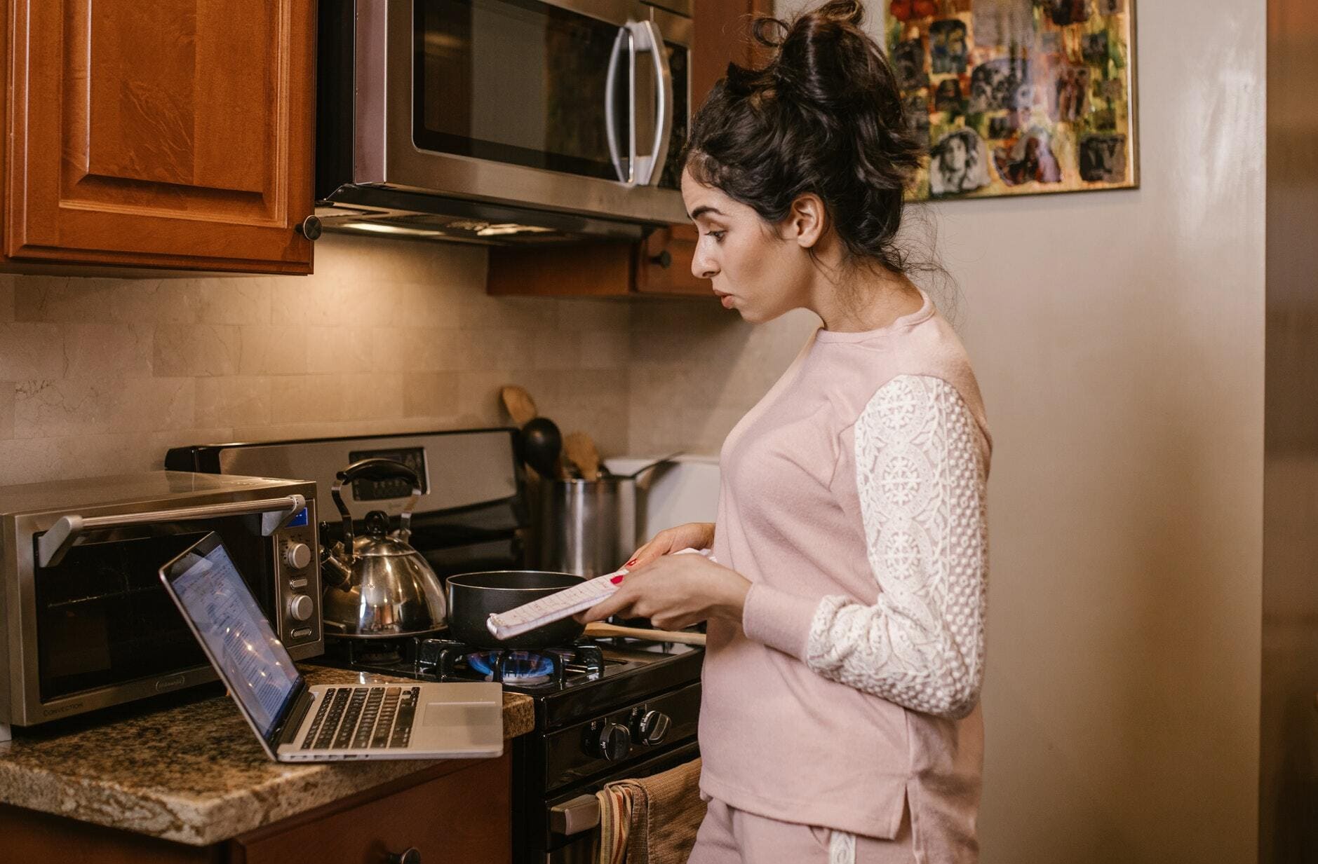 Ernährung Home-Office Eine Frau im pastellfarbenen Hausanzug steht vor dem Herd und schaut konzentriert in ihren Laptop auf der Arbeitsplatte. In der Hand hält sie einige Unterlagen.