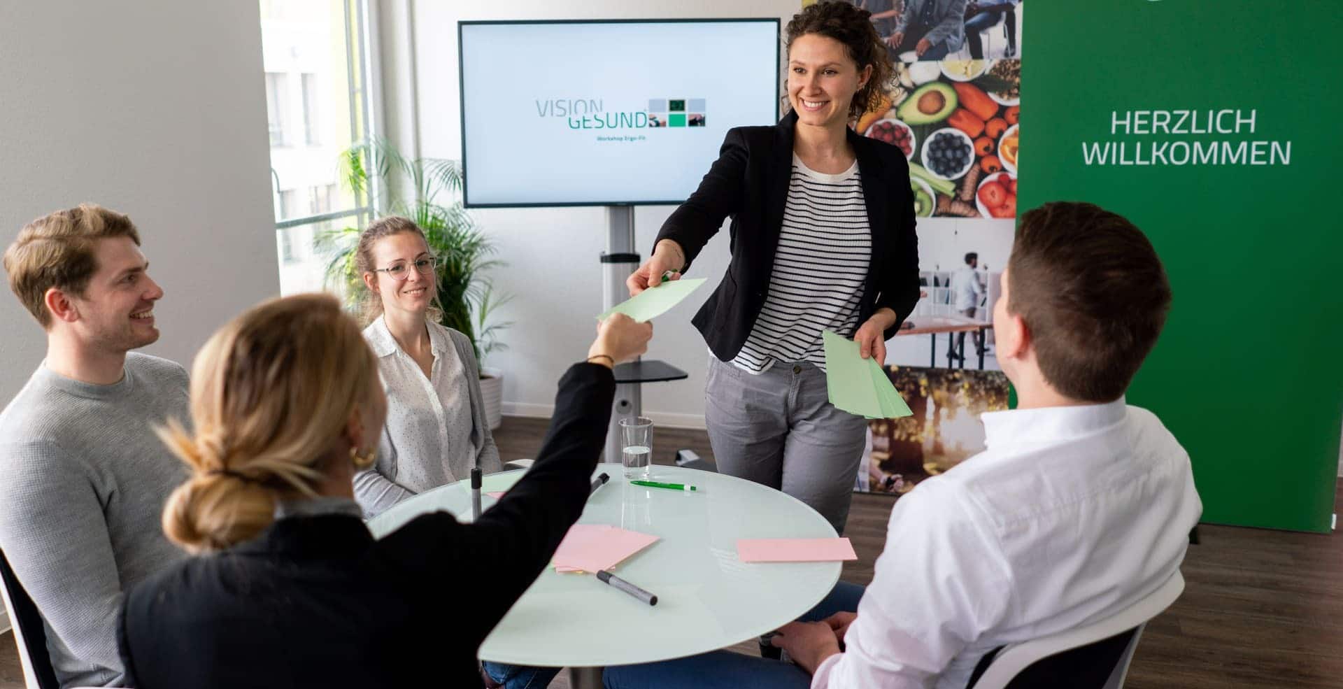 Die stehende VisionGesund-Expertin reicht einer Teilnehmerin der am Tisch sitzenden, vierköpfigen Arbeitsgruppe lächelnd einen bunten Zettel. Im Hintergrund sind Aufsteller des Unternehmens VisionGesund in dessen Farben Silber und Grün zu sehen. Die Stimmung wirkt entspannt, es herrscht eine angenehme Atmosphäre.