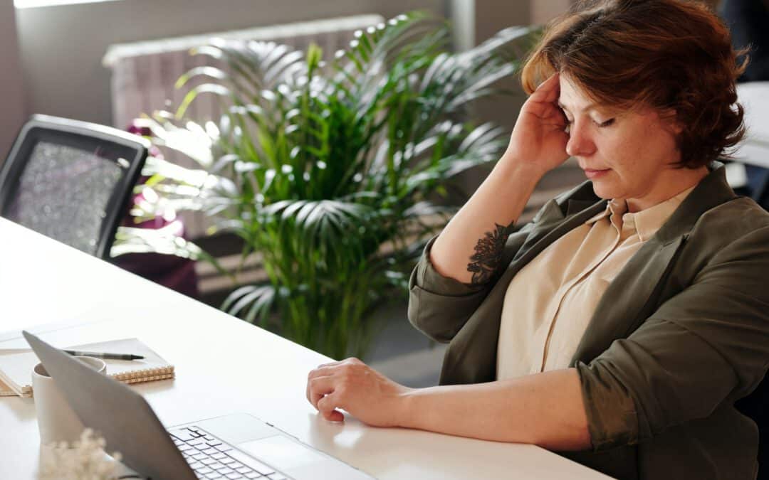 Frau sitzt am Schreibtisch und leidet unter Long-COVID Symptomen und hält sich den Kopfn