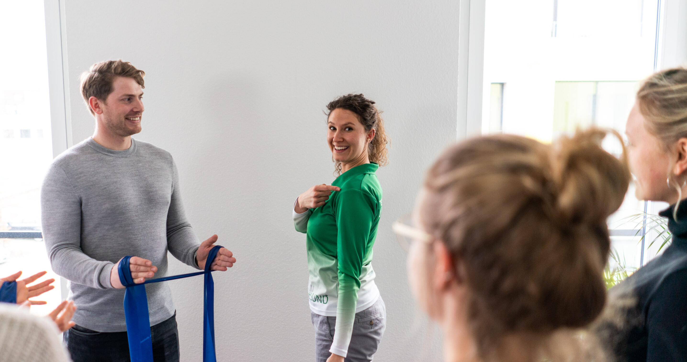 Mehrere Erwachsene stehen in einem hellen Raum und machen gemeinsam Übungen mit Resistancebändern. Eine Person zeigt die Bewegung, während die anderen aufmerksam zuschauen und mitmachen.