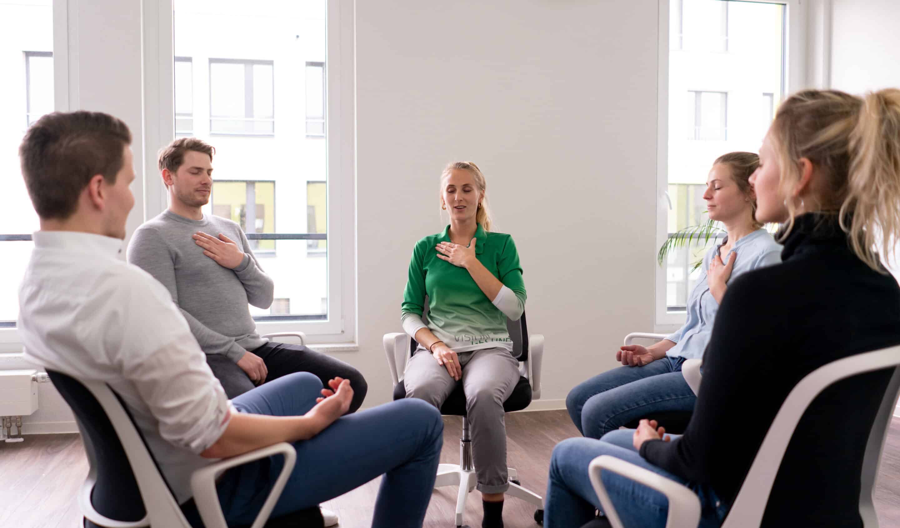 Eine Gruppe von fünf Personen sitzt im Kreis in einem hellen Raum. Die Frau in der Mitte trägt grüne Sportkleidung und führt eine Atem- oder Entspannungsübung vor, indem sie eine Hand auf die Brust legt. Die Teilnehmenden sitzen entspannt auf Stühlen und folgen ihrer Anleitung.