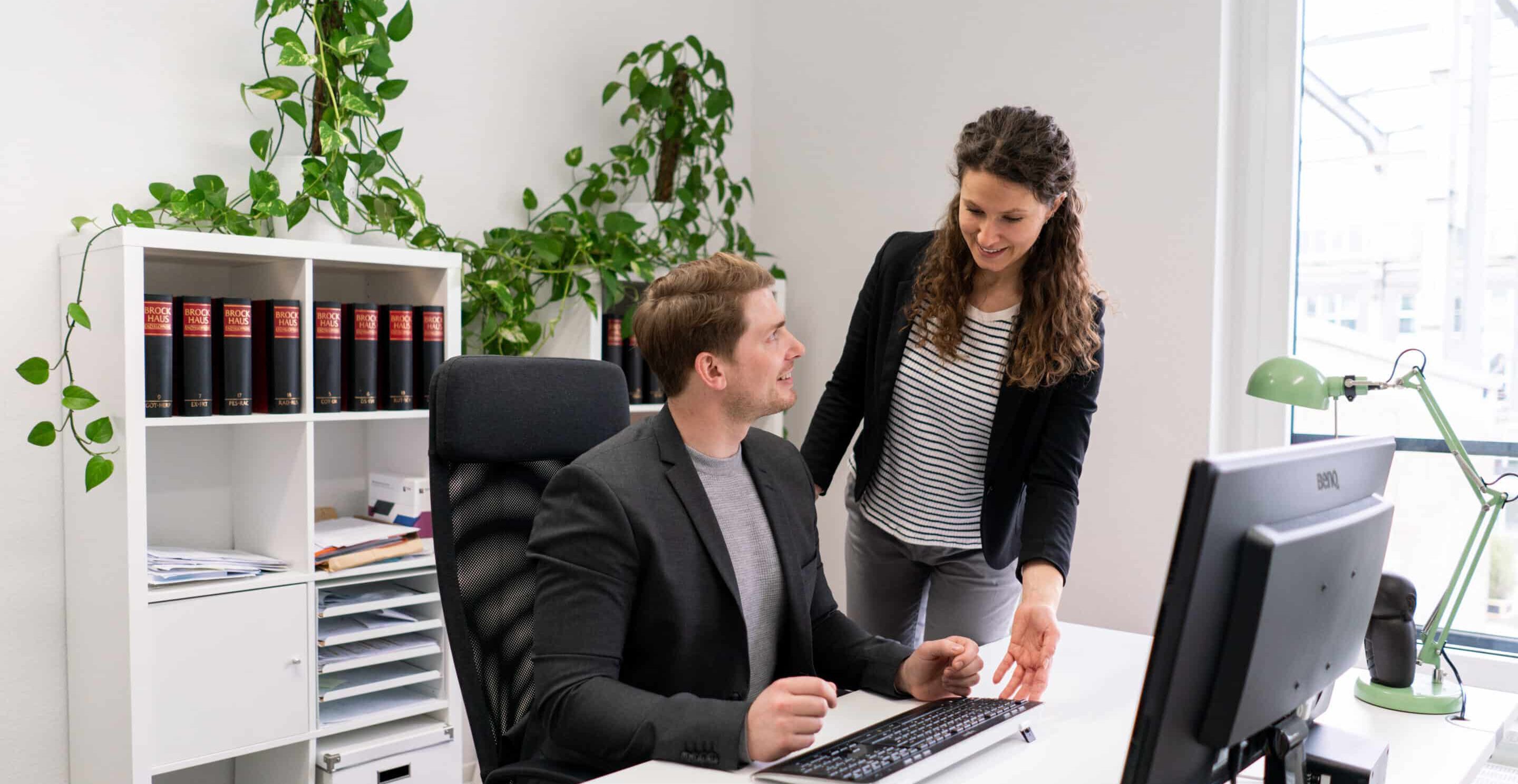 Zwei Personen arbeiten gemeinsam am Computer im Büro, eine gibt Tipps für eine gesunde und gute Arbeitshaltung. Das Bild zeigt Betriebliche Gesundheitsförderung am Arbeitsplatz.