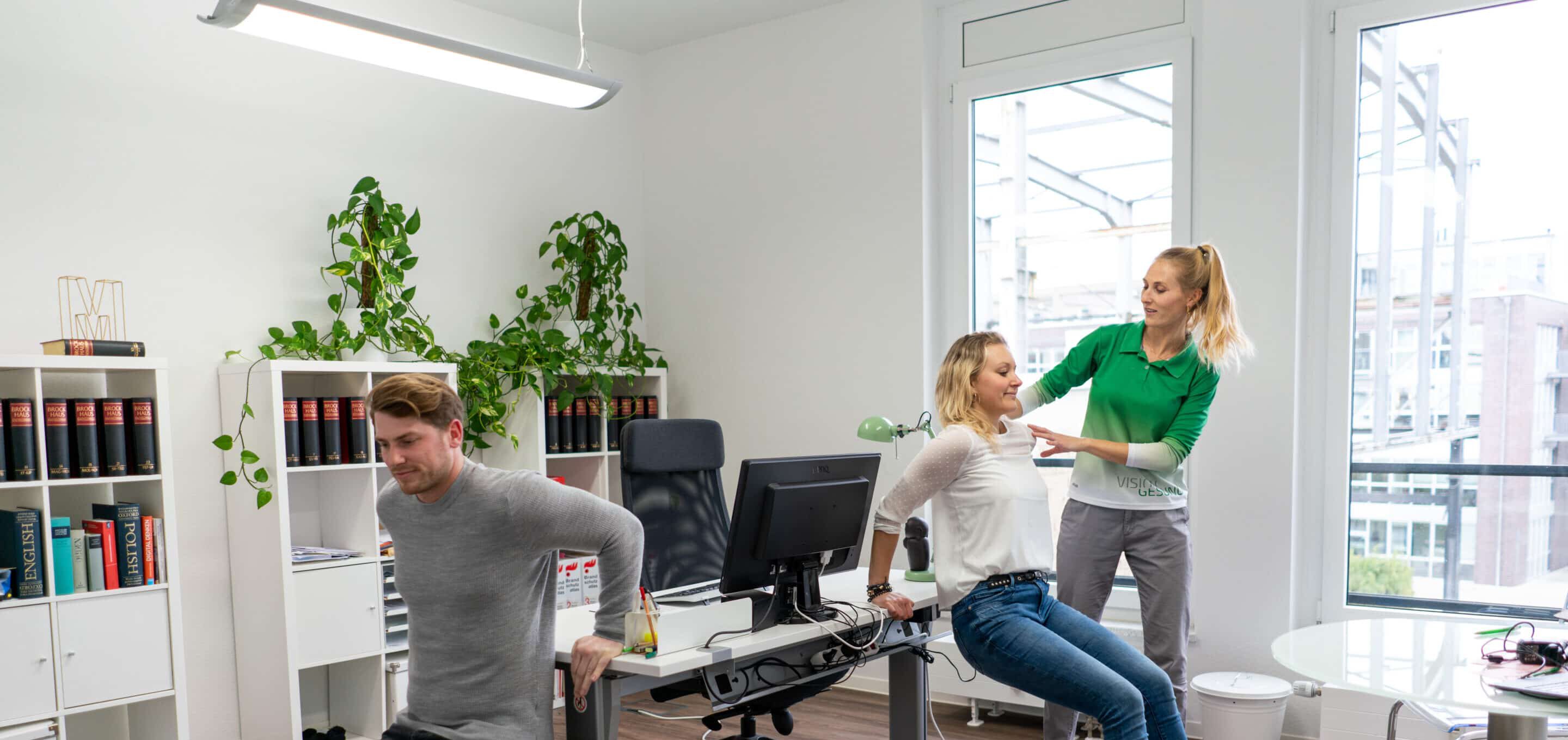 Eine Gruppe befindet sich in einem Büro und führt Übungen oder Demonstrationen zu ergonomisch korrektem Arbeiten durch. Der Raum ist modern und lichtdurchflutet.