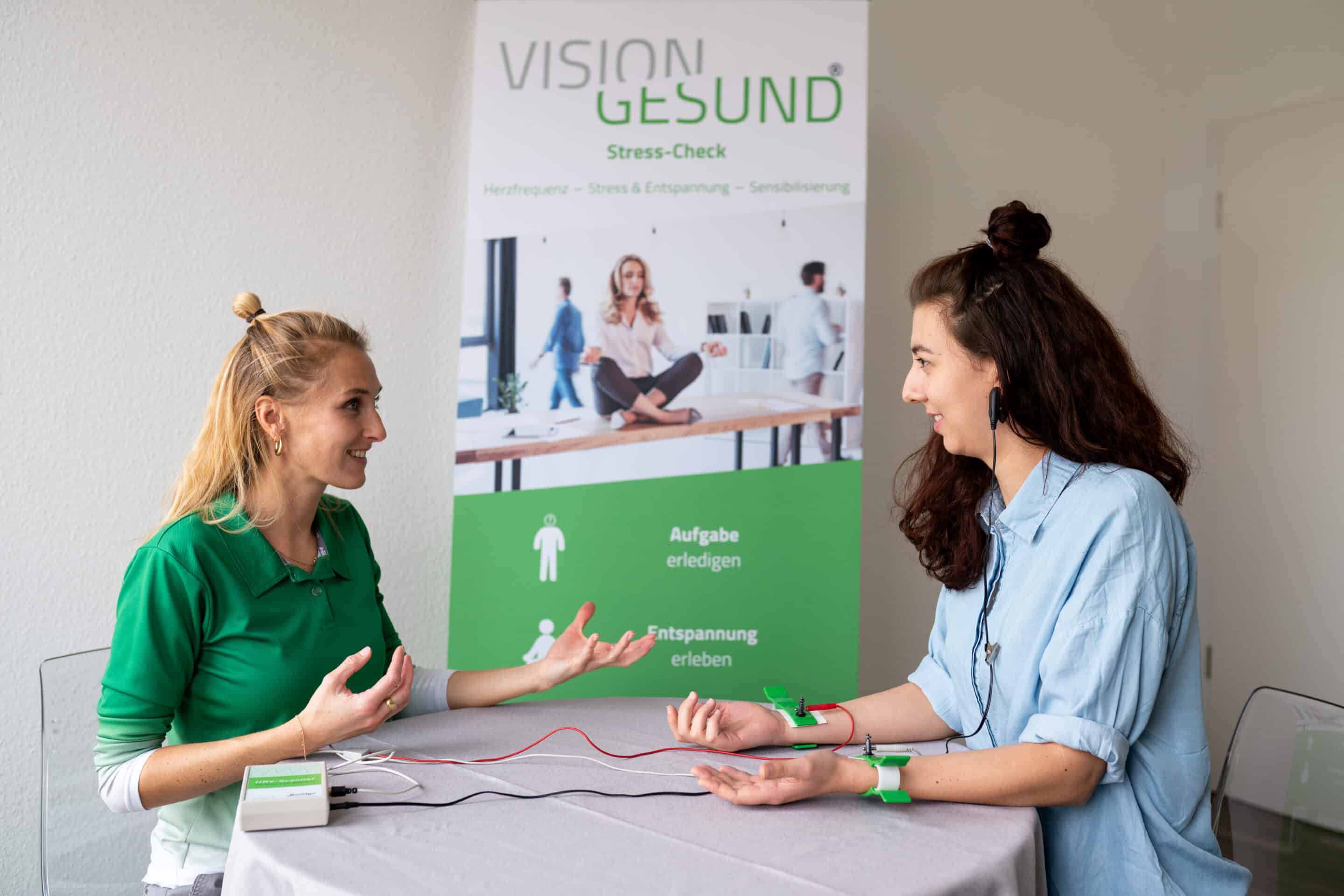 Stress-Check Zwei Frauen sitzen sich an einem Tisch gegenüber und führen einen Stress-Check durch. Beide tragen Sensoren an den Handgelenken, während eine Mitarbeiterin von Vision Gesund den Ablauf erklärt. Im Hintergrund steht ein Roll-up mit der Aufschrift ‚VISION GESUND – Stress-Check‘ und Bildern zu Stress- und Entspannungsmessung.