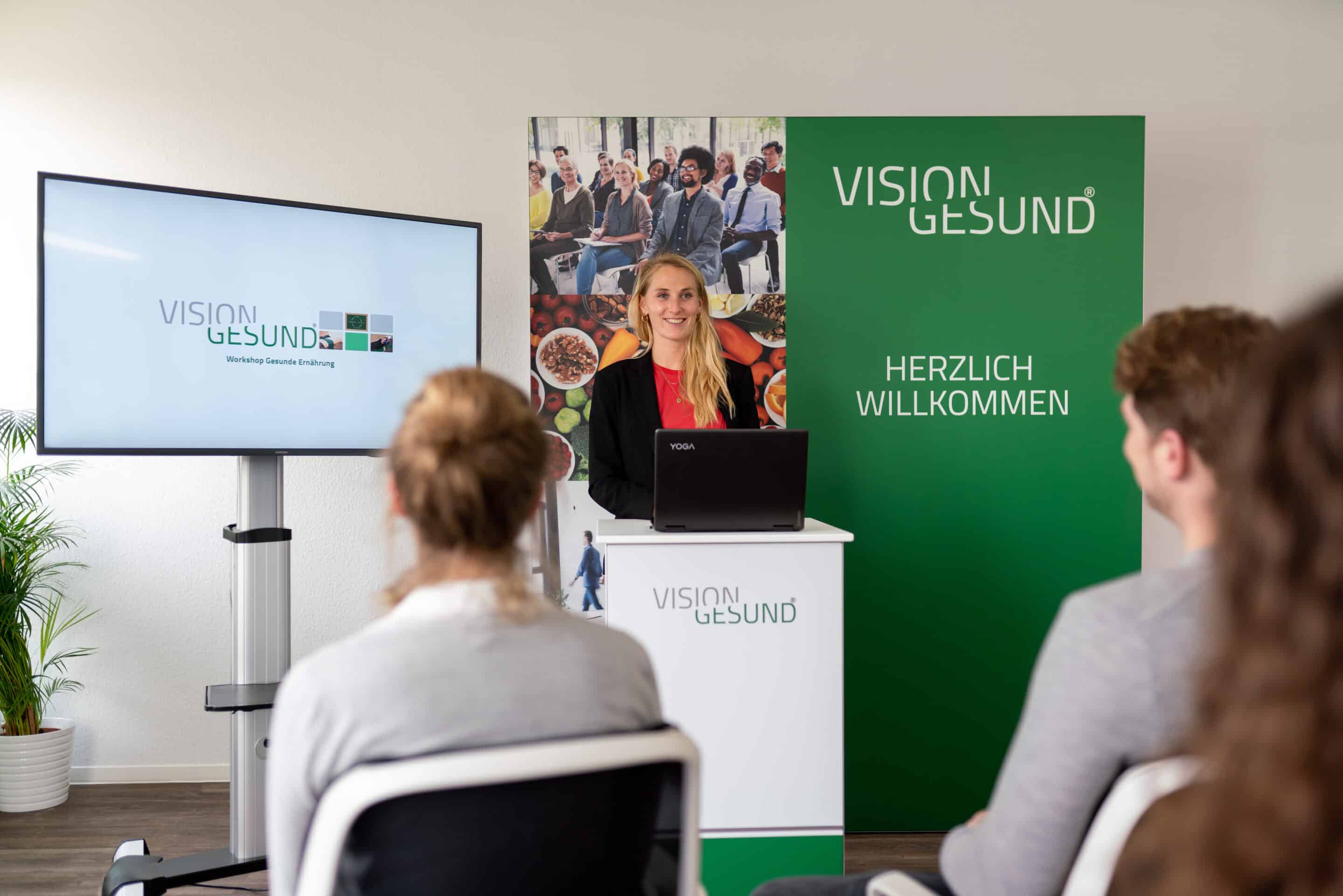 Body-Mind-Connection Eine Referentin von Vision Gesund steht hinter einem Rednerpult und begrüßt Teilnehmende in einem Seminarraum. Vor ihr sitzen mehrere Personen und hören zu. Links zeigt ein Monitor das Vision‑Gesund‑Logo, rechts steht ein grünes Roll‑up mit der Aufschrift ‚Herzlich Willkommen‘.