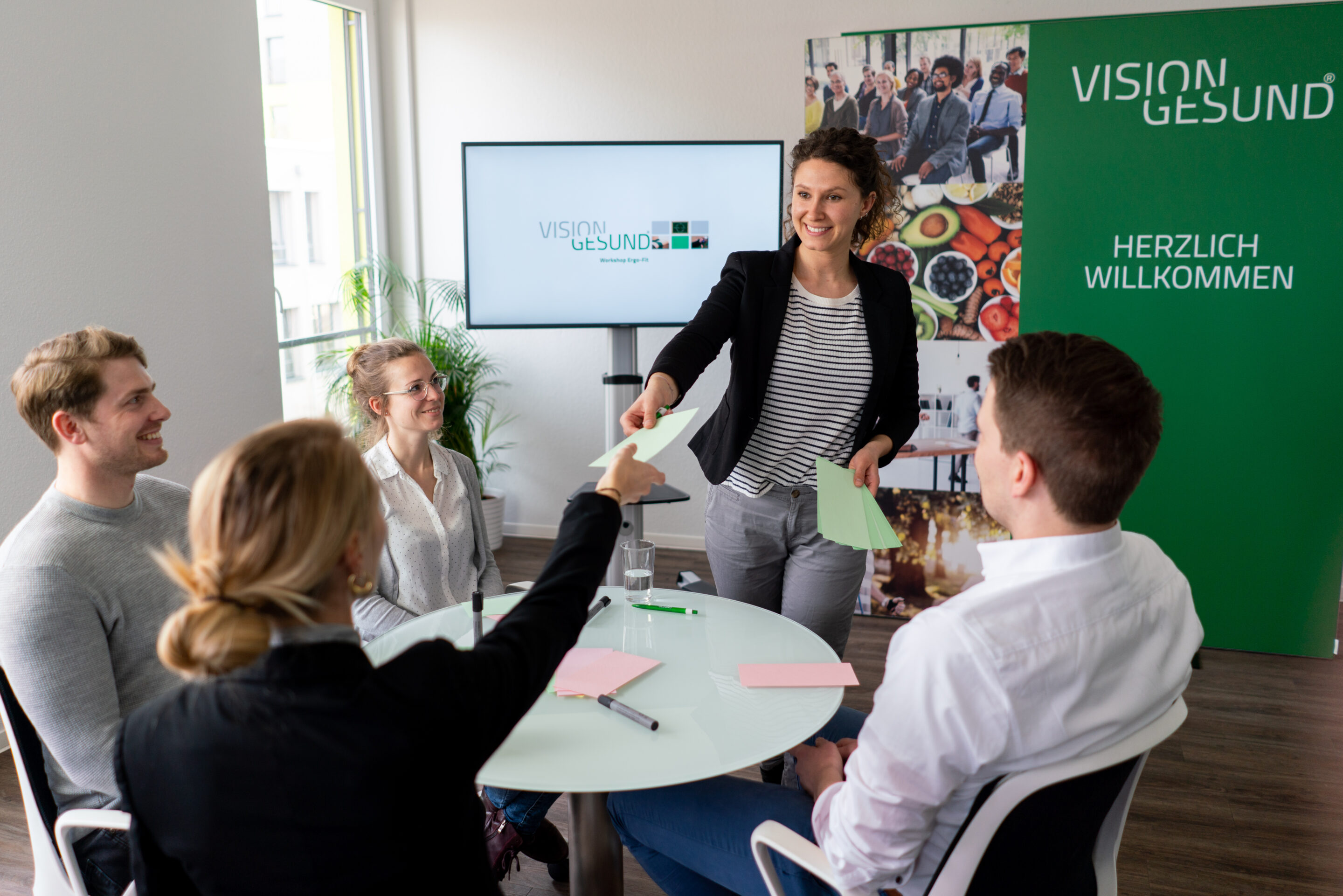 Mitarbeitende sitzen in einem Meetingraum, während eine Fachkraft das BGF‑Programm „Vision Gesund“ vorstellt.