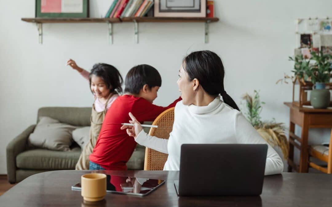 Mutter dreht sich zu ihren im Hintergrund spielenden Kindern, während sie am Laptop von zuhause aus arbeitet. Eine typische Situation im Home-Office
