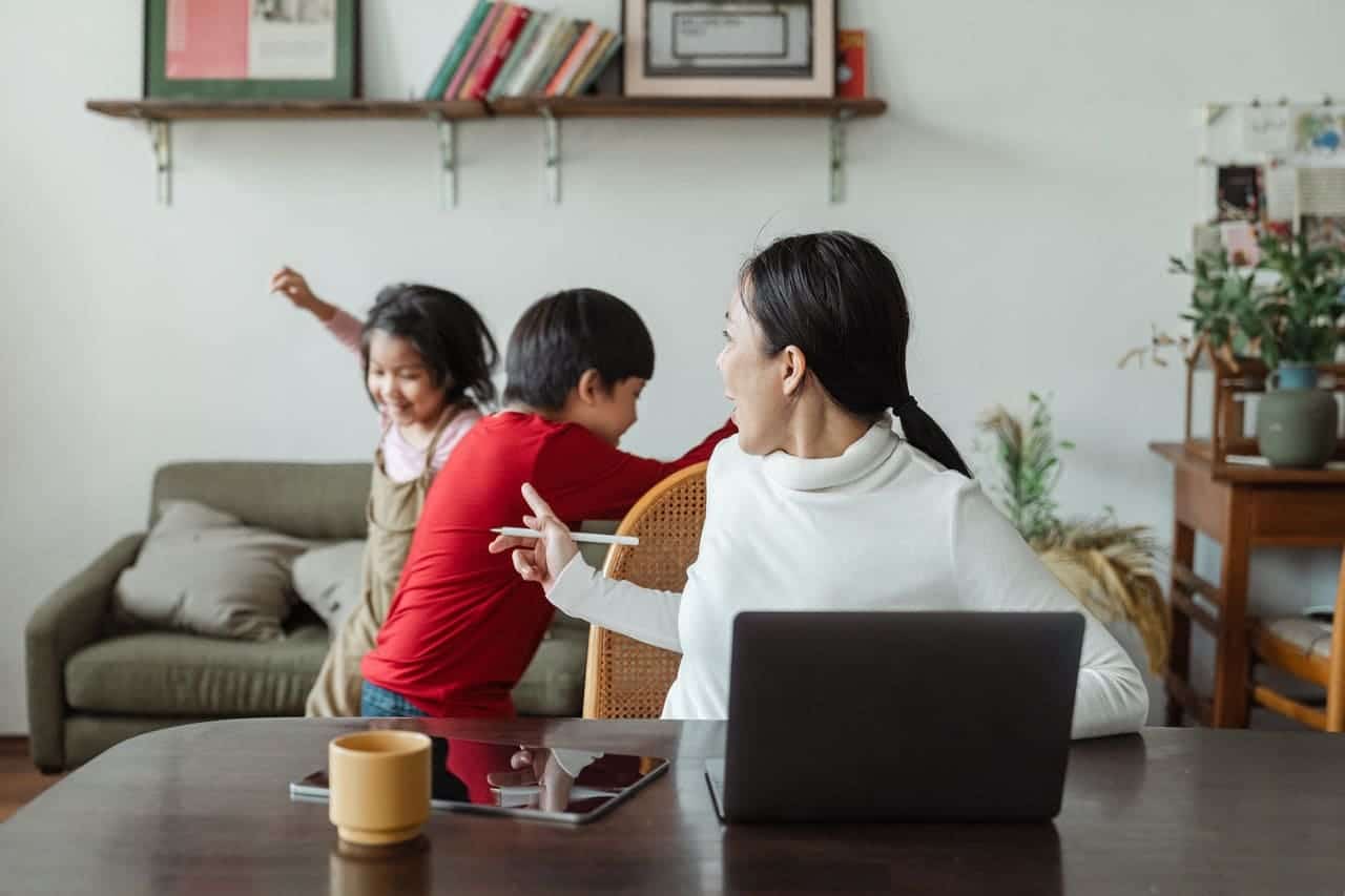 Psyche Home-Office Mutter dreht sich zu ihren im Hintergrund spielenden Kindern, während sie am Laptop von zuhause aus arbeitet. Eine typische Situation im Home-Office