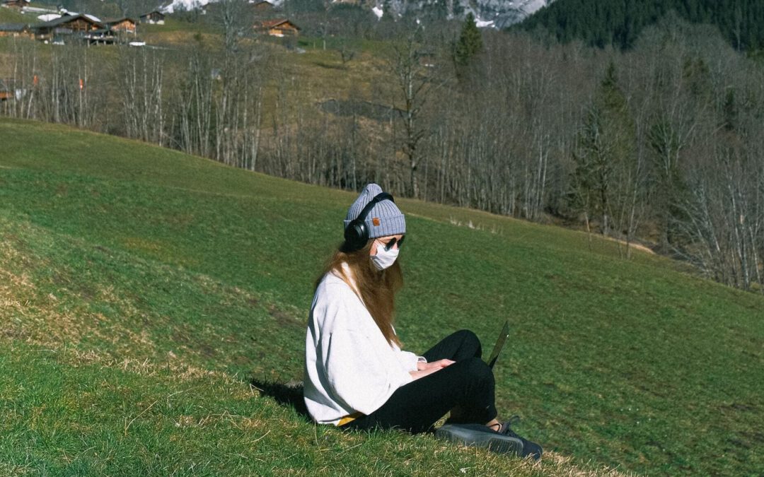 Eine junge Frau sitzt mit Maske und Mütze bekleidet im Schneidersitz auf dem Abhang einer Bergwiese. Sie trägt Kopfhörer und schaut auf den Bildschirm des Laptops auf ihrem Schoß. Im Hintergrund sind Ansätze des Gebirges und Almen zu erkennen.