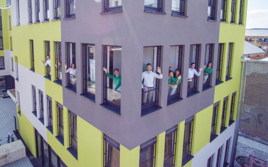 Auf dem Bild ist das gelb-graue Gebäude des VisionGesund-Büros zu sehen. Aus den Fenstern des mehrstöckigen Hauses winken aus der zweiten Etage die Mitarbeitenden des VisionGesund-Teams.