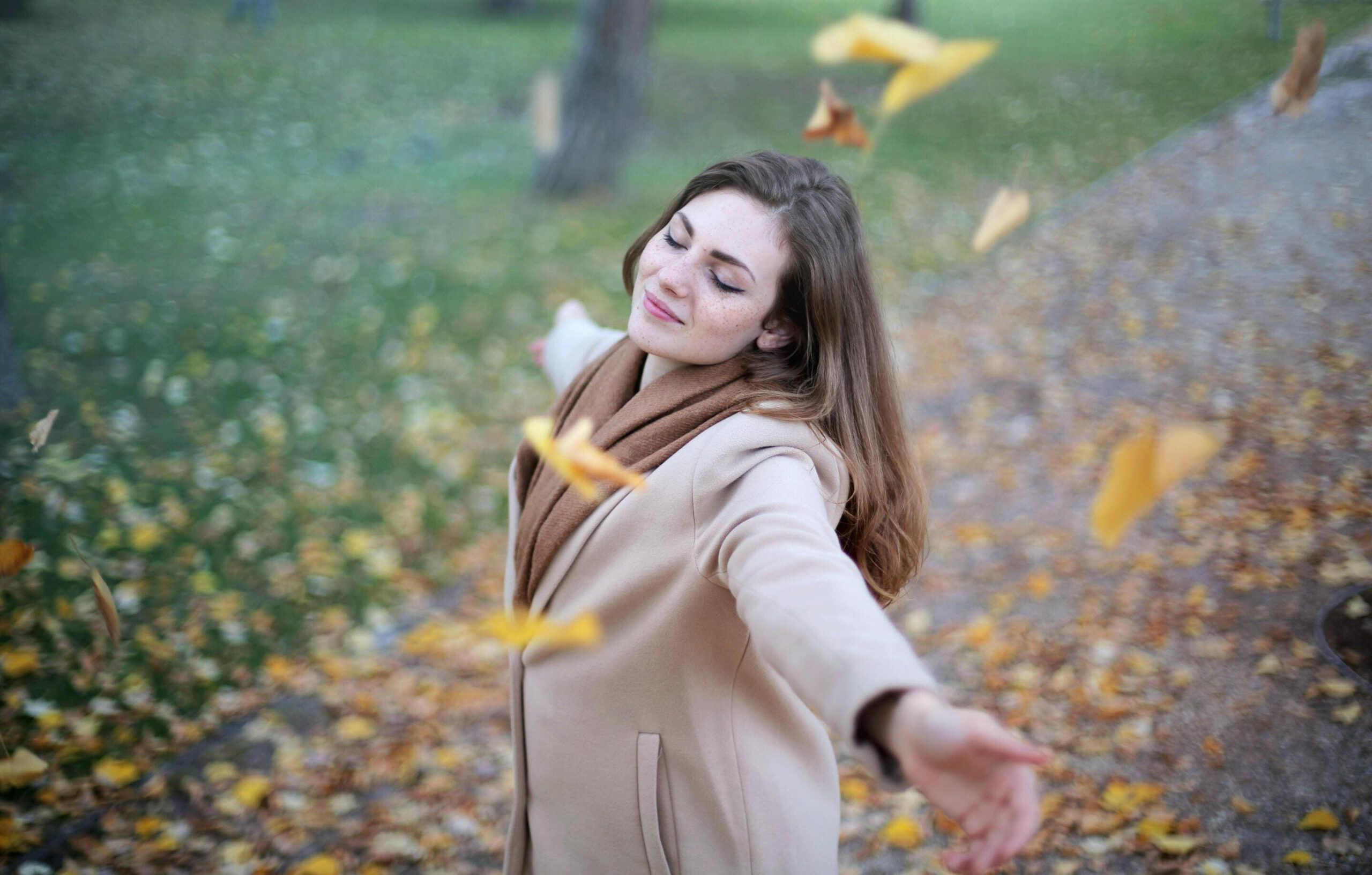 Achtsamkeit Zentral im Bild ist eine Frau mit langen braunen Haaren und die Arme zur Seite ausgestreckt, zu sehen. Sie trägt einen beigen Wintermantel und einen braunen Schal. Ihre Augen sind geschlossen, der Kopf leicht nach oben geneigt und gelbe Blätter fallen von oben in das Bild hinein.