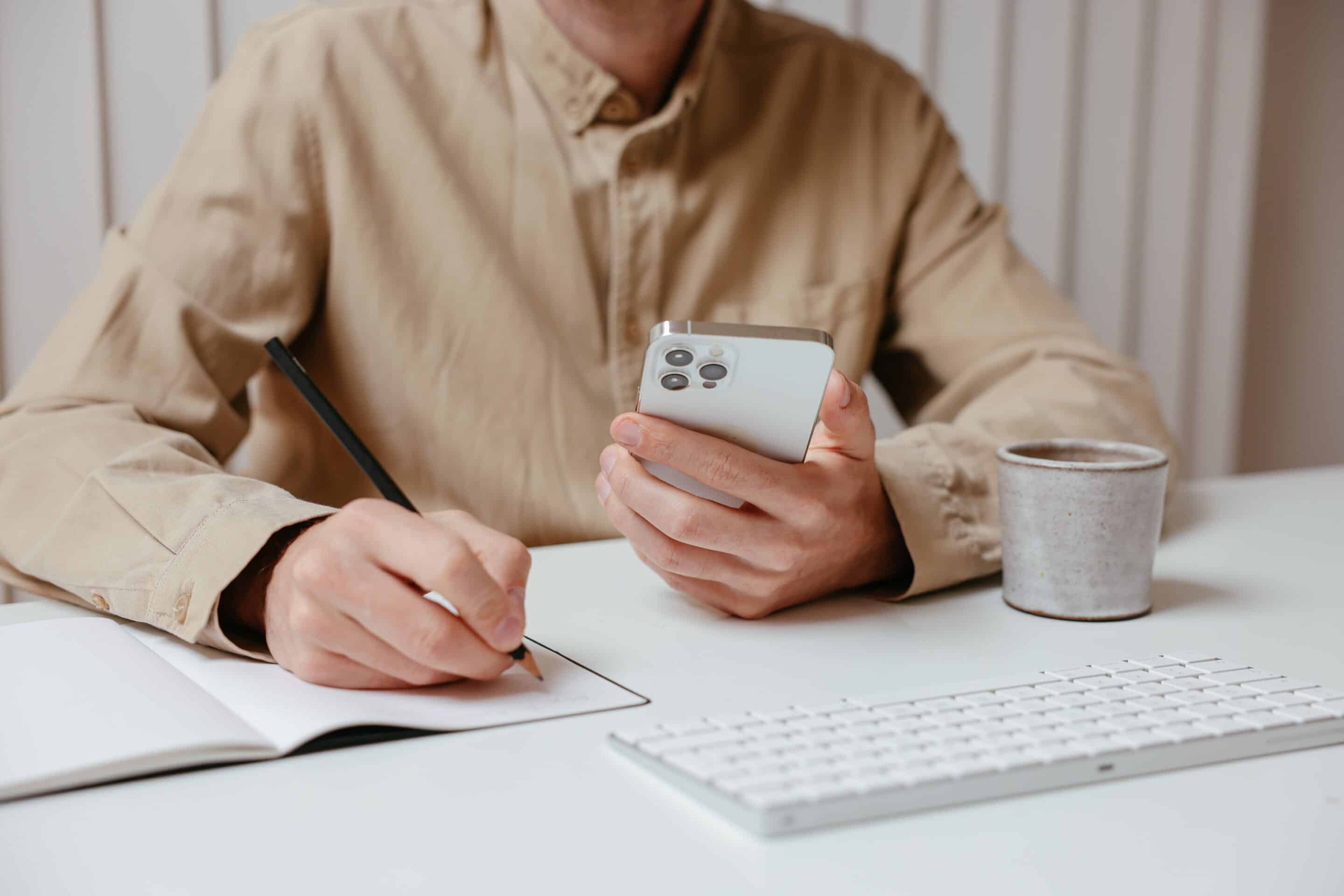 pexels-cup-of-couple-7657730 Mann hält ein Smartphone am Arbeitsplatz fest.