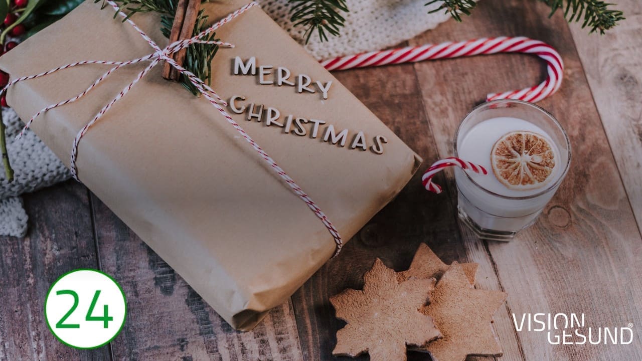 Frohe Weihnachten Heiligabend Auf der linken Seite des Bildes liegt ein in Paketpapier geschnürtes Päckchen, um das viele weihnachtliche Dekors wie eine rotweiße Zuckerstange in einem weißen Getränk, Holzsterne, Zimtstangen und Tannenzweige drapiert sind. Den Untergrund bildet ein stilvoller Holztisch. Auf dem Paket prangen die Worte: Merry Christmas.