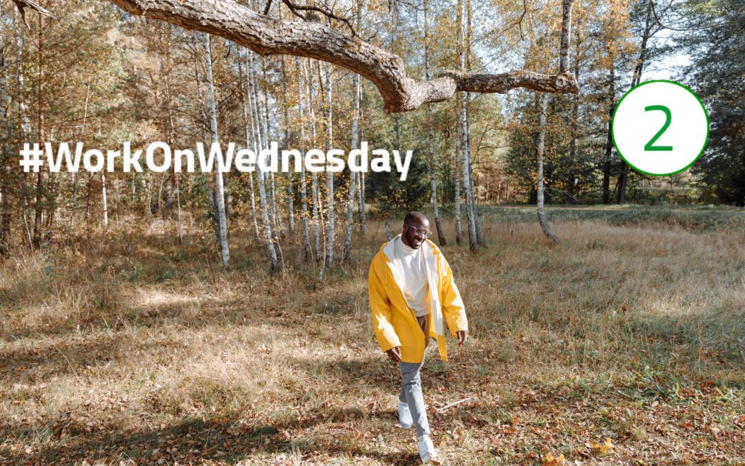 Ein Mann mit offener, gelber Regenjacke spaziert durch den Wald und trägt ein Lächeln im Gesicht. Links im Bild steht "#WorkOnWednesday" geschrieben und oben rechts im Bild ist die Zahl "2" zu sehen.